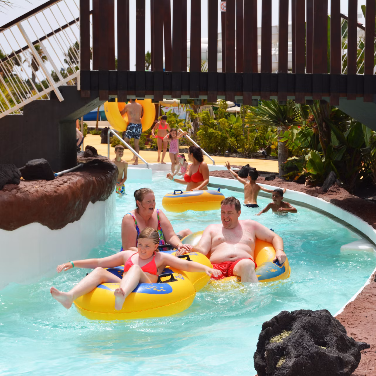 Adults and children riding inflatable tubes on a lazy river in a water park, with lush greenery and other people in the background.