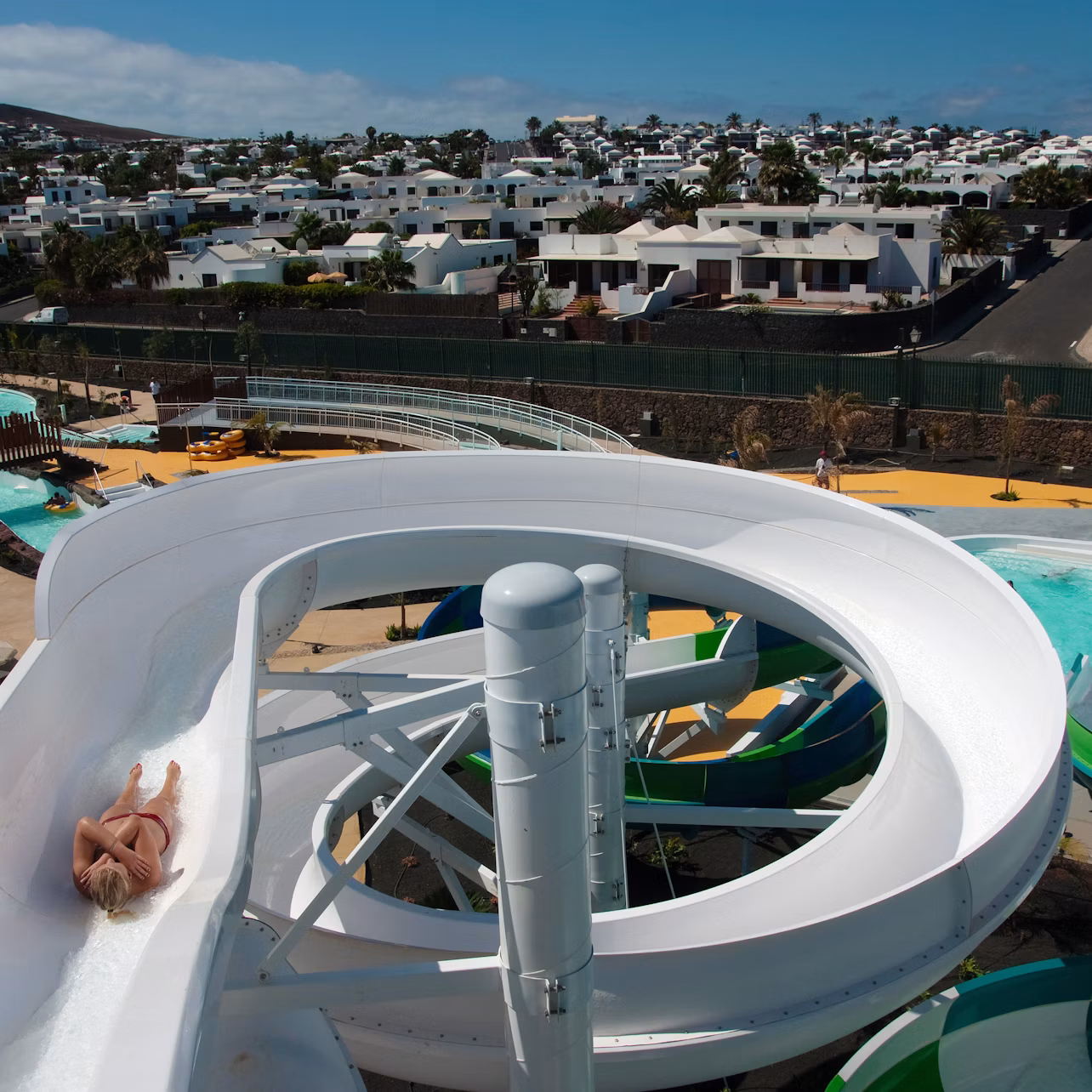 A person sliding down a white twisty water slide at a water park with rows of white houses in the background.