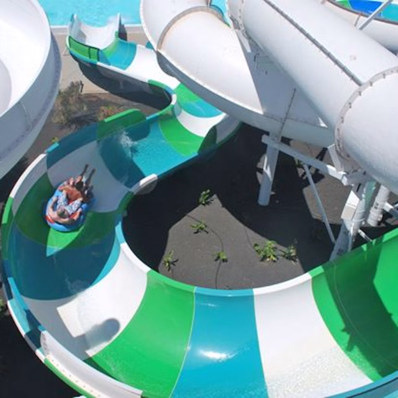 People on a raft slide down a colorful water slide, surrounded by larger white tube slides, with a pool in the background.