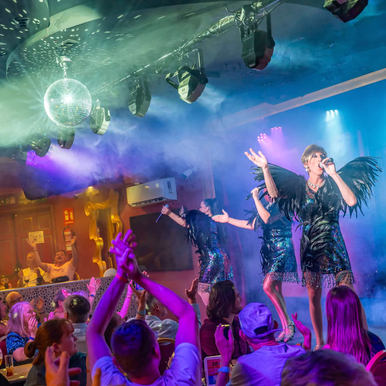 Three performers in matching black outfits sing on stage under a disco ball, with a cheering audience in a lively atmosphere.