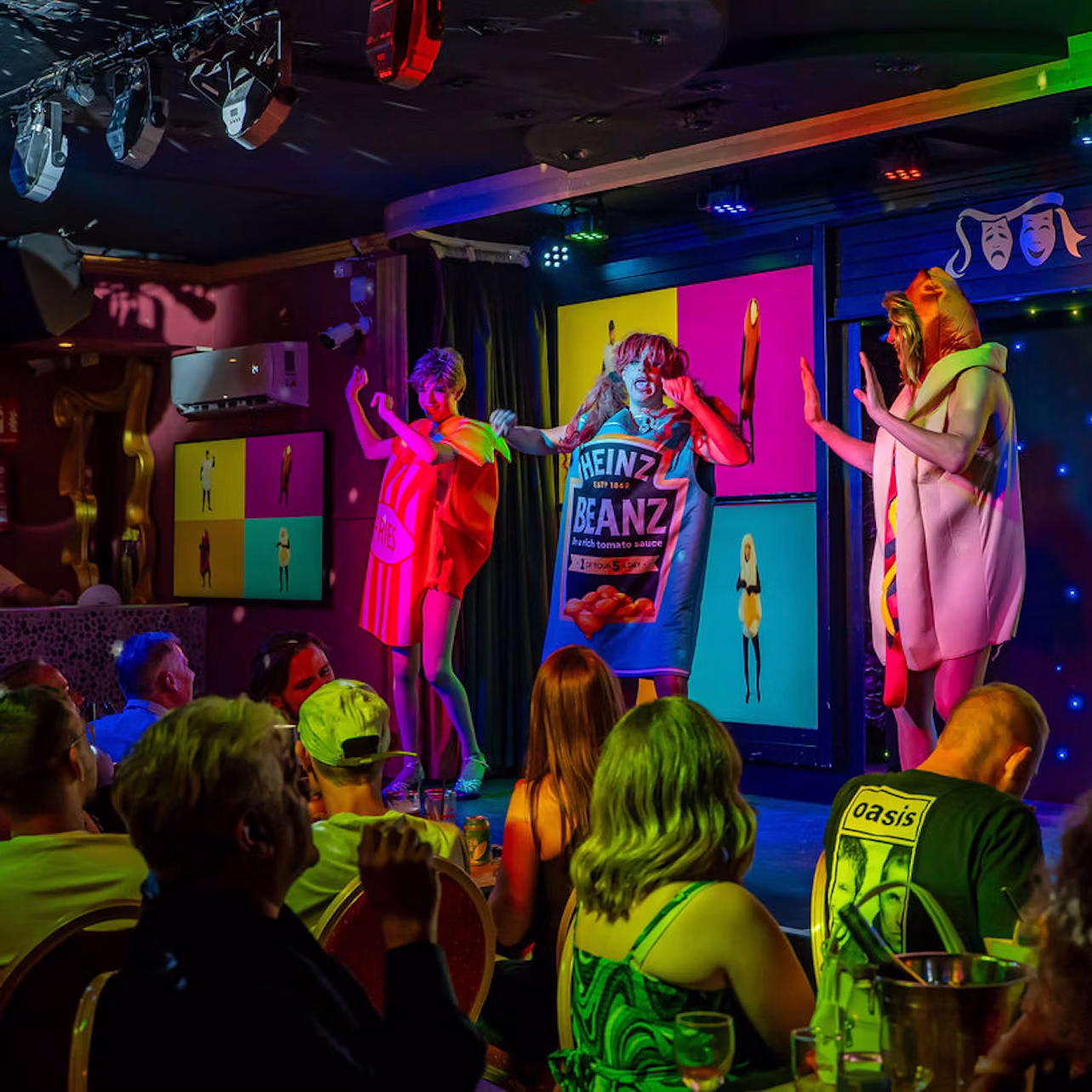 Three performers in colorful costumes, including food-themed outfits, entertain an audience in a vibrant stage setting.