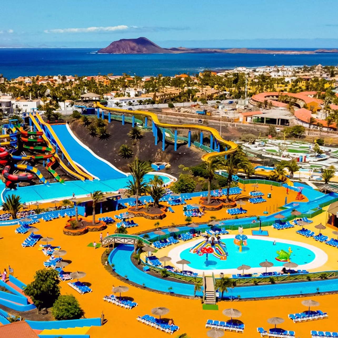 A colorful water park with pools, slides, and lounging areas, set against a coastal town and ocean backdrop.