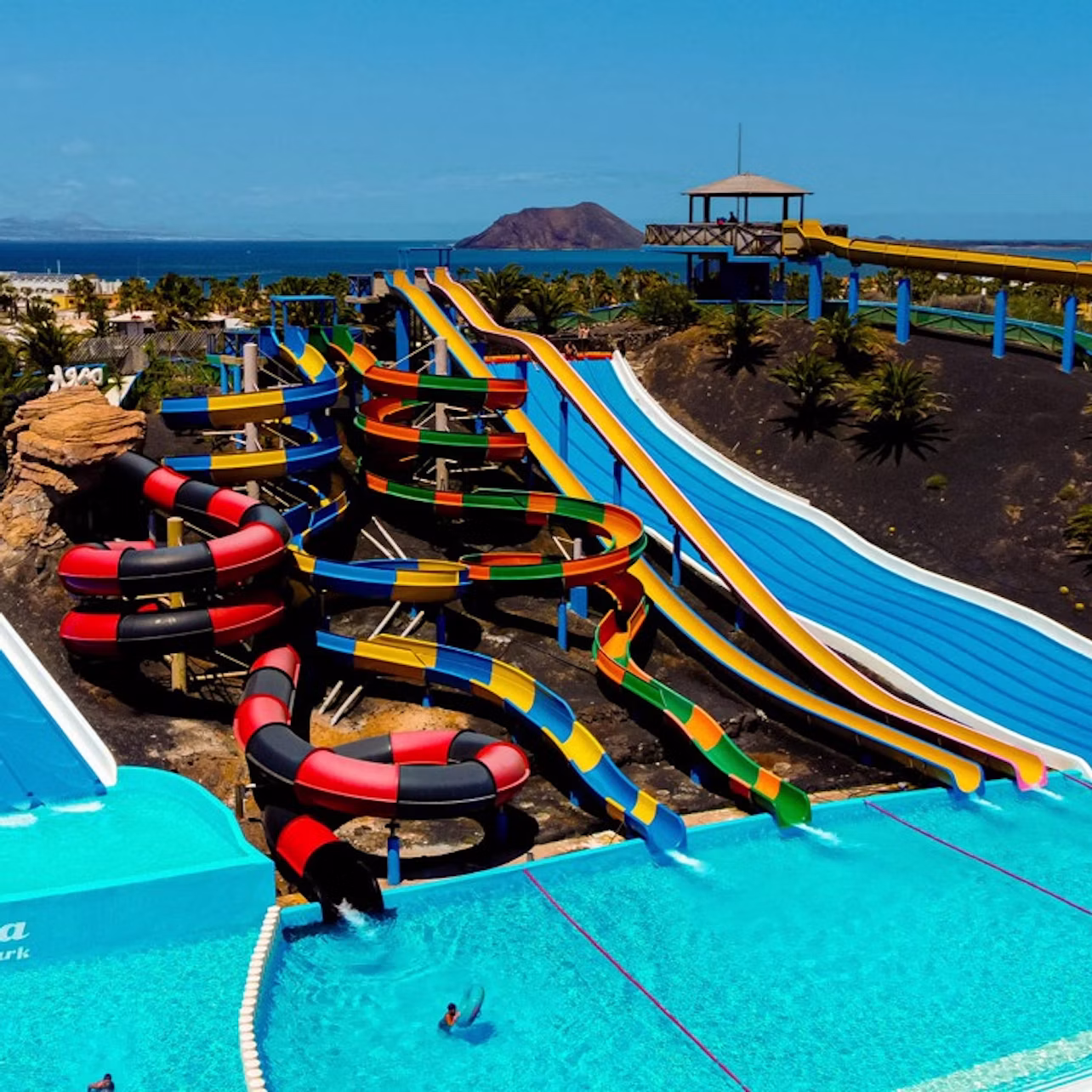 A colorful water park with multiple slides leading into pools, surrounded by a scenic landscape under a clear blue sky.