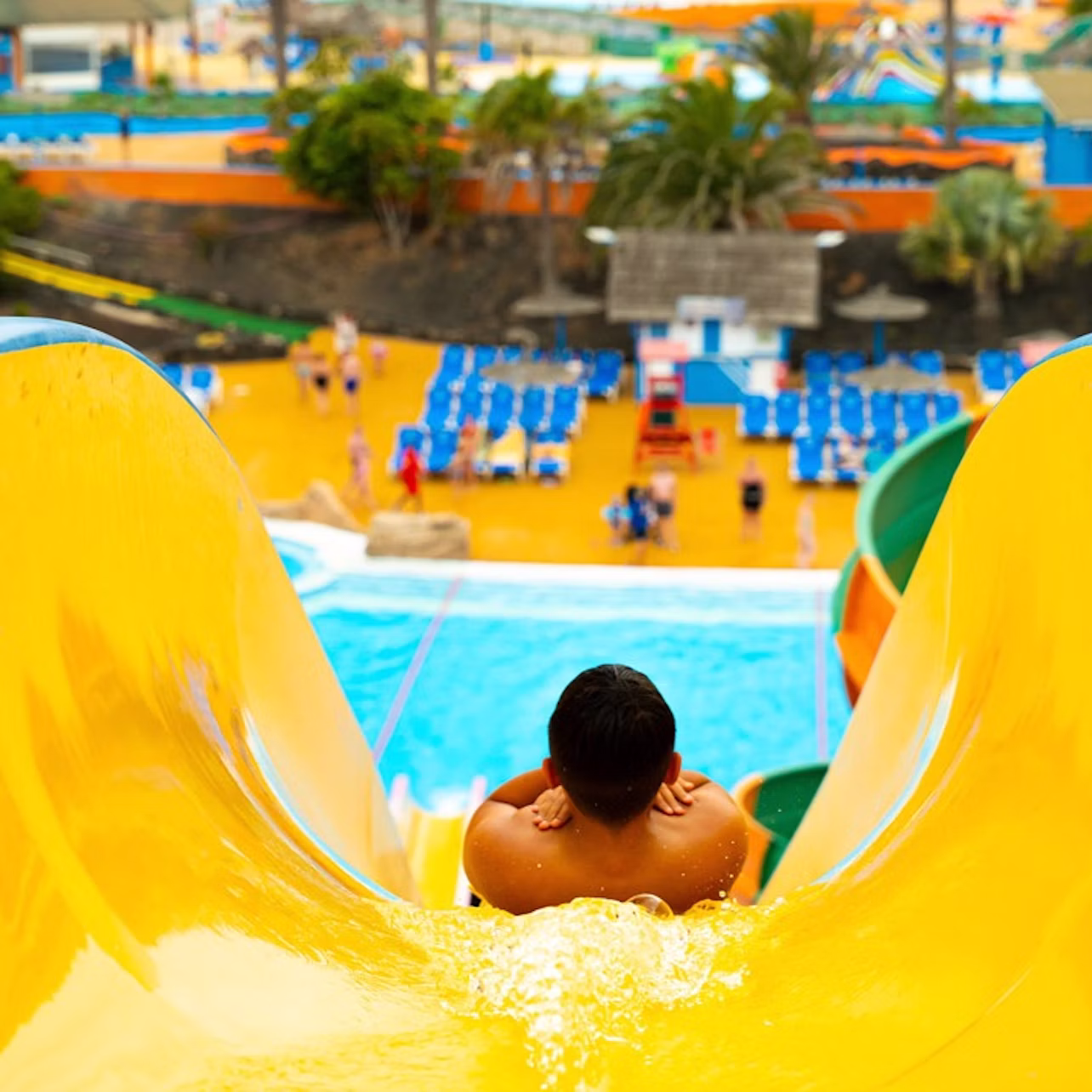 A person sliding down a yellow water slide at a water park, with pools, chairs, and palm trees in the background.