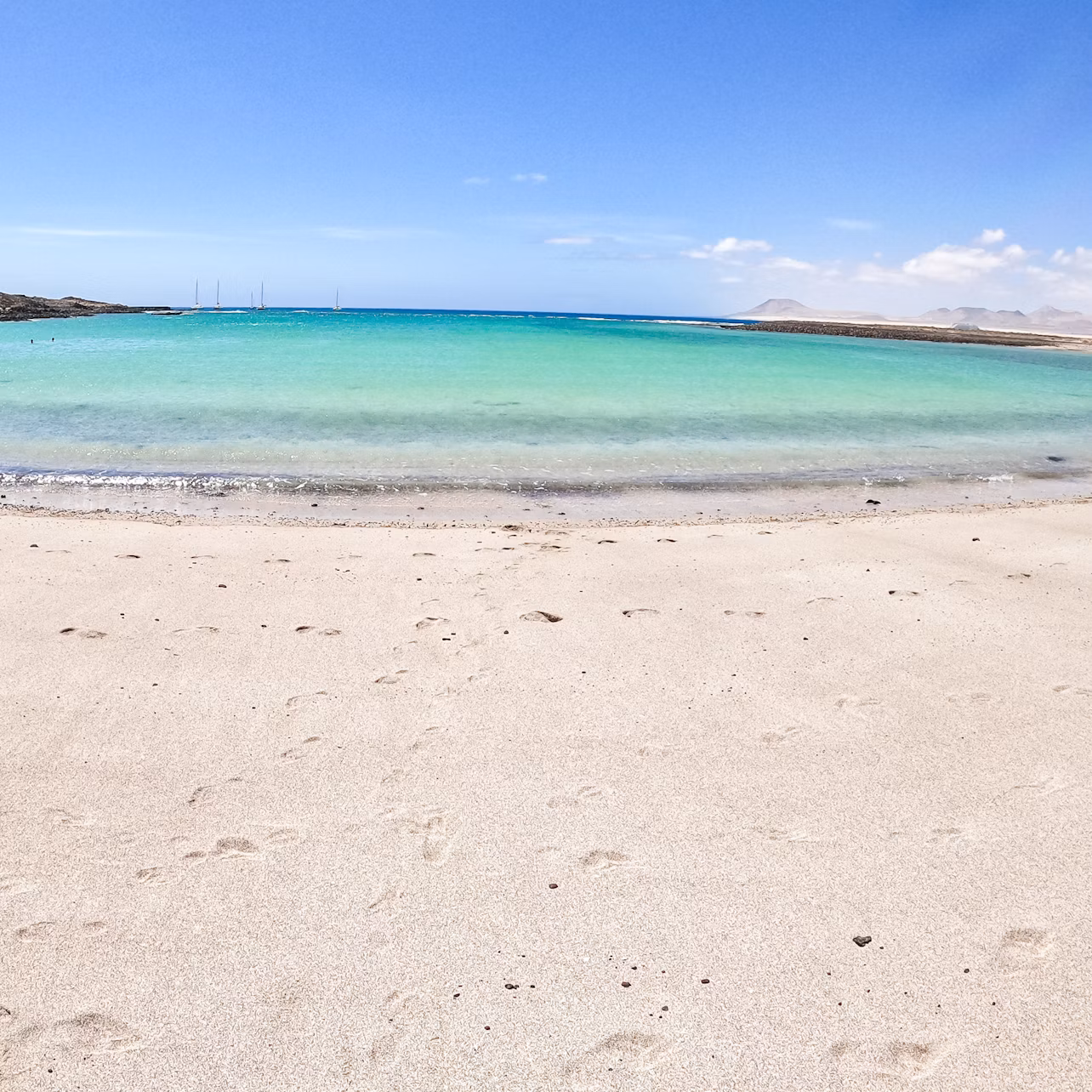 A serene beach with smooth sand, clear turquoise water, and a bright blue sky with light clouds.