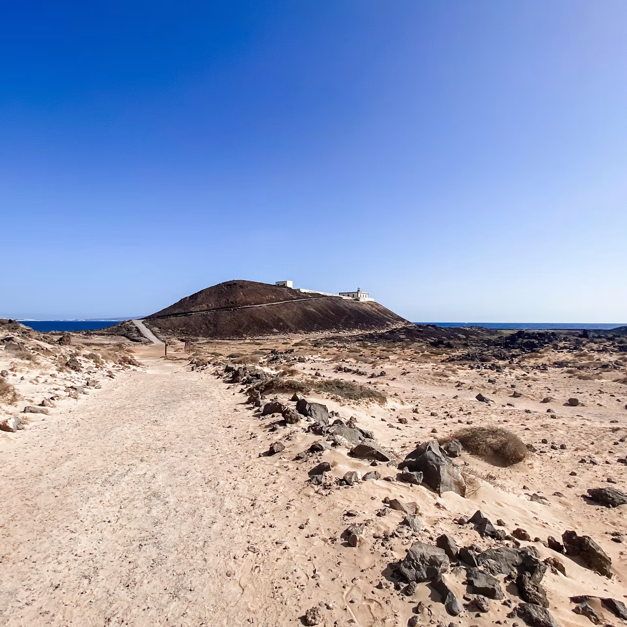 A rocky dirt path leads to a hill with a structure on top, set under a clear blue sky and surrounded by sparse desert terrain.