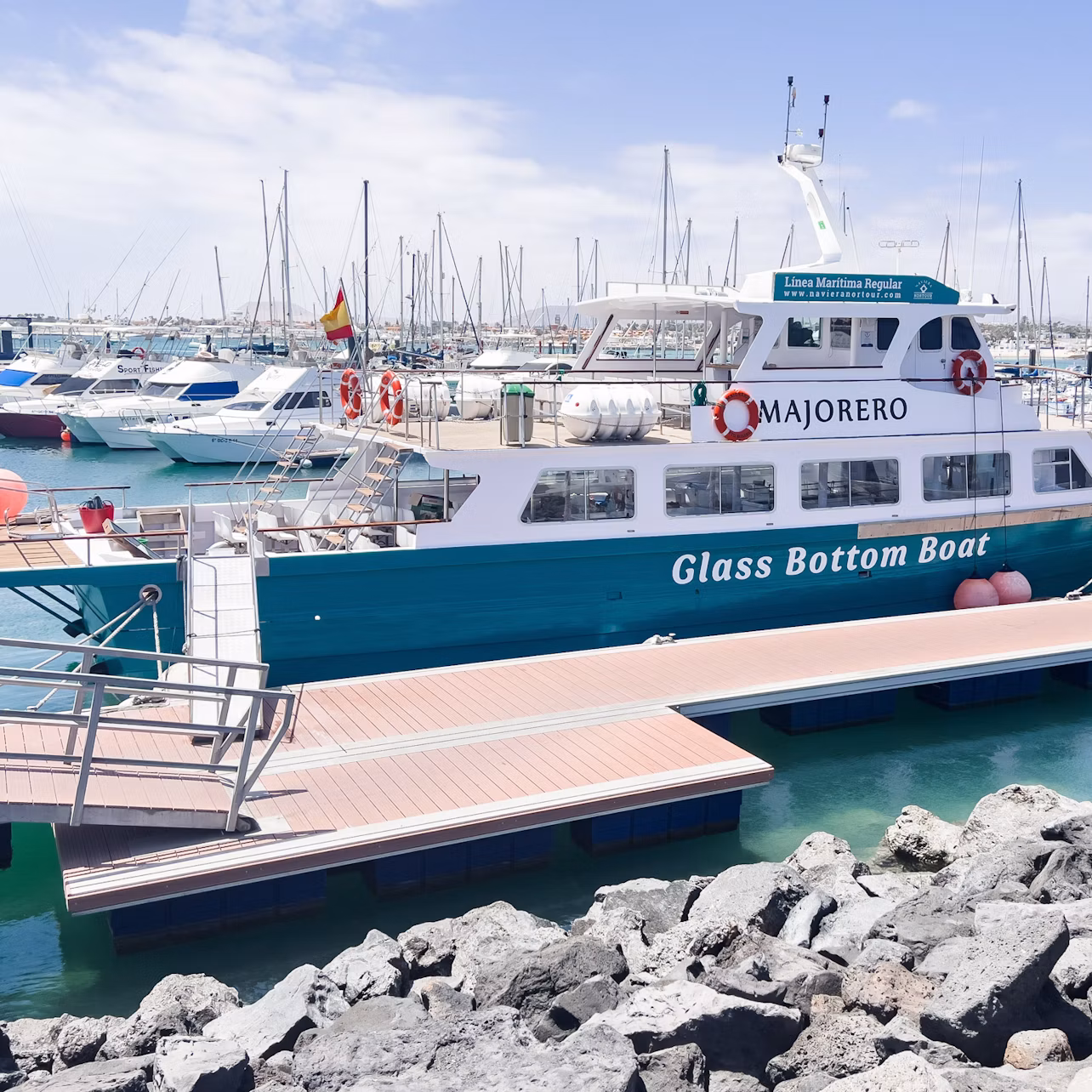 A turquoise and white glass-bottom boat docked at a marina with other boats on a sunny day. Rocks in the foreground.