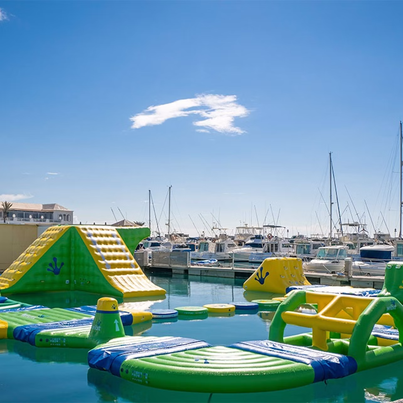 Inflatable water park with slides and obstacles floats on water, with a marina full of boats in the background under a clear sky.