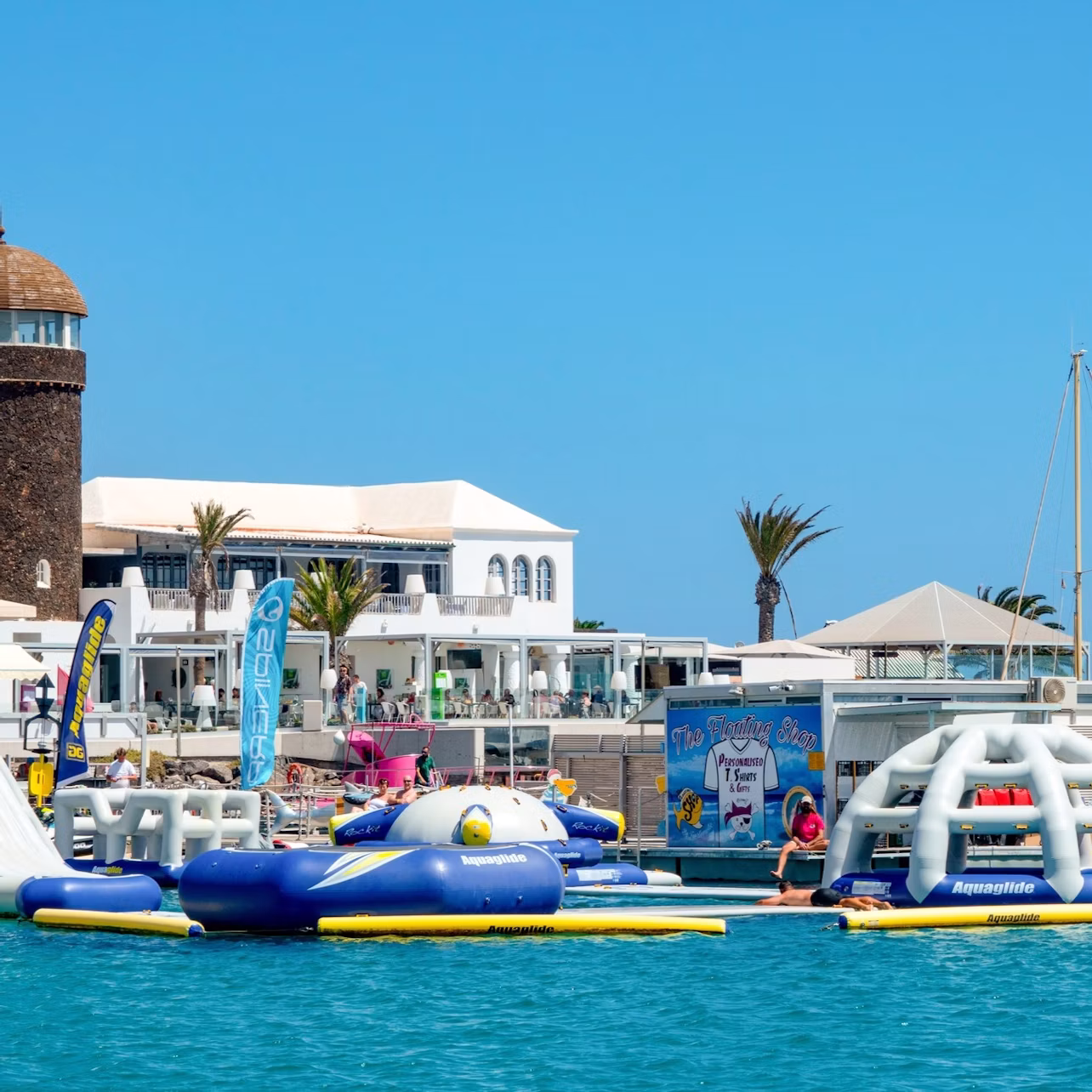 Floating water park with inflatable structures in front of a coastal building and palm trees under a bright blue sky.