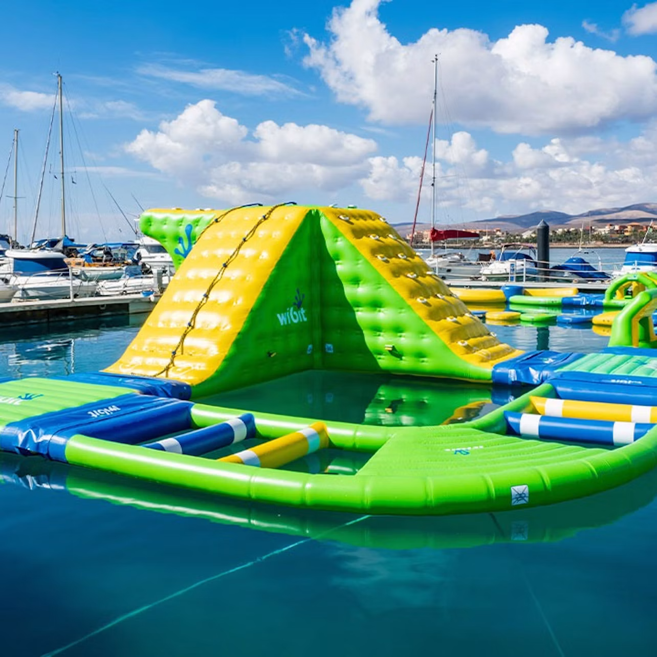 Inflatable water obstacle course with yellow and green slide, floating in a marina with docked boats and mountains in the background.