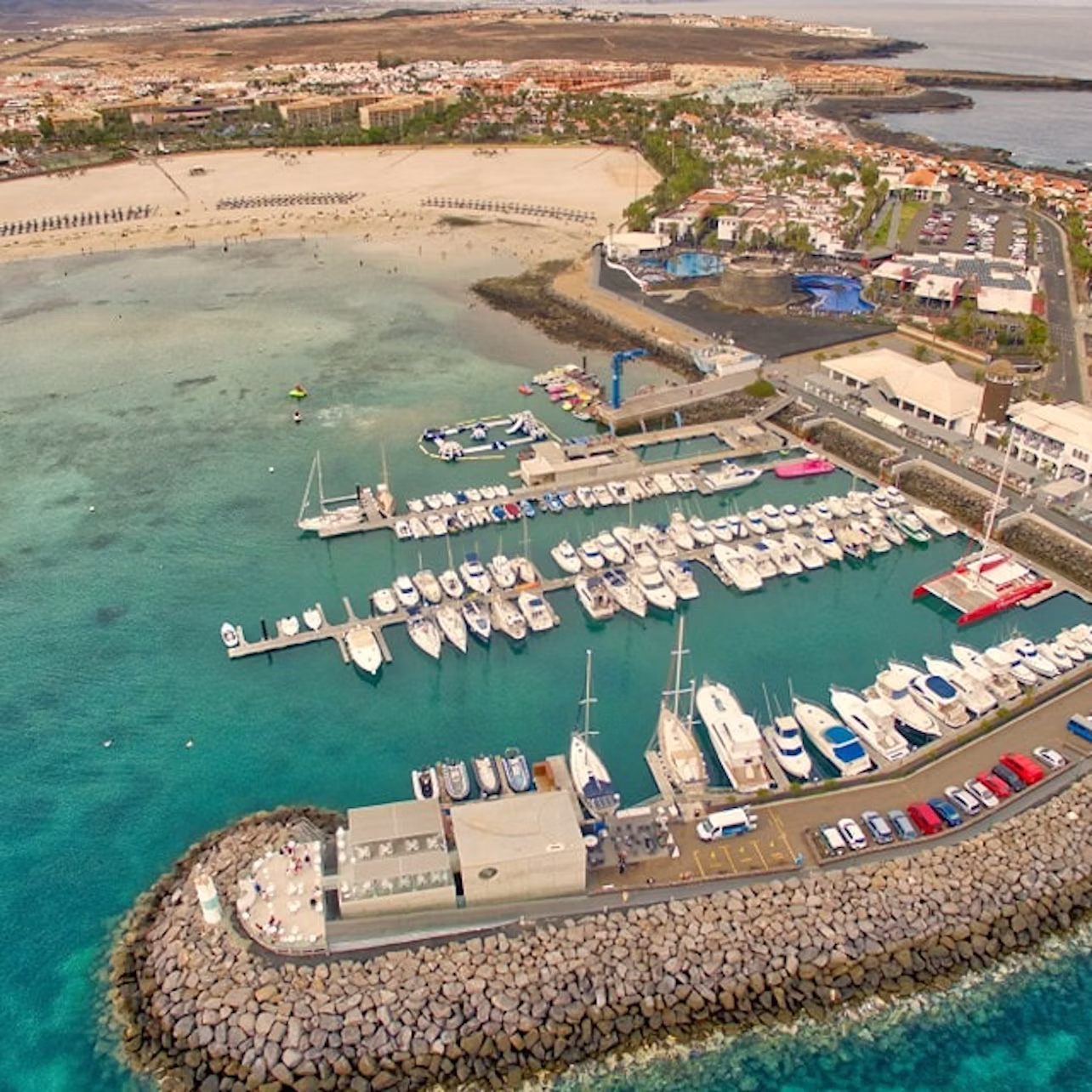 Aerial view of a marina with numerous boats docked, adjacent to a coastal town with a beach and various buildings.