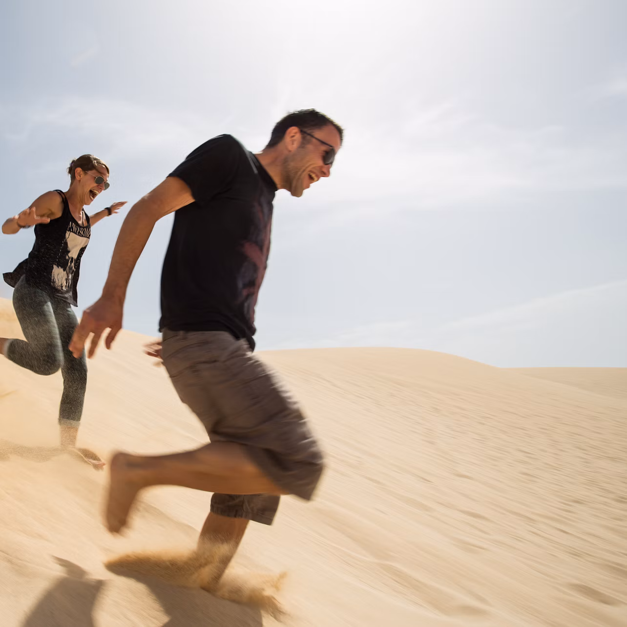Two people gleefully running down a sunlit sand dune under a clear sky.