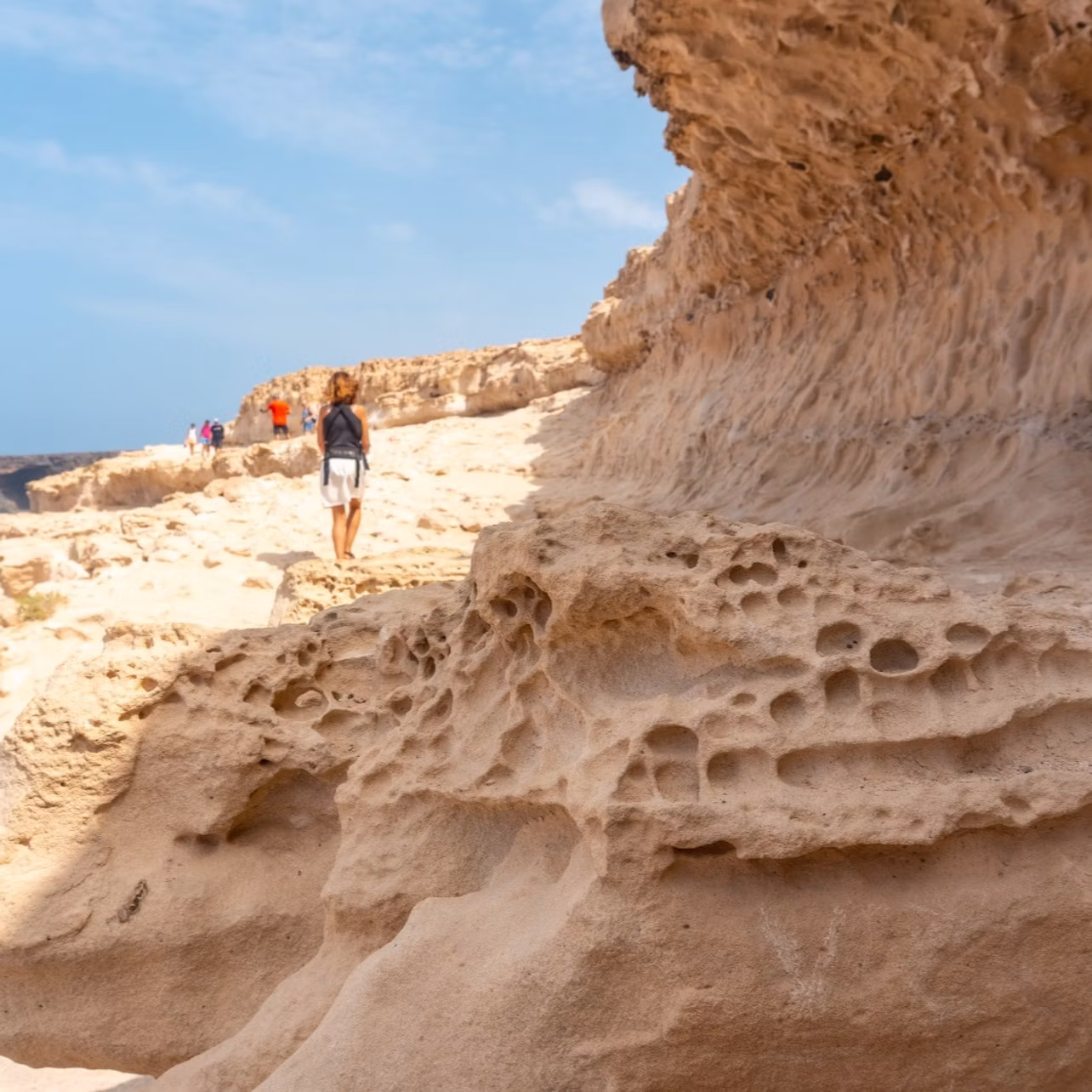 A person wearing a backpack walks along a rocky, eroded cliff path under a clear blue sky, with others in the distance.