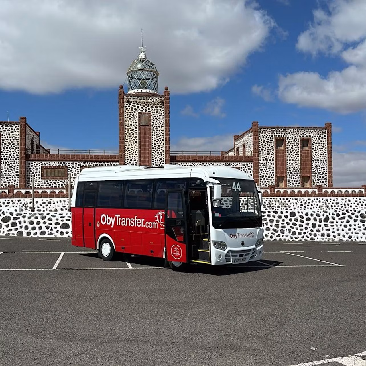 Red and white tour bus parked in front of a stone building with a tower and clock, under a partly cloudy sky.