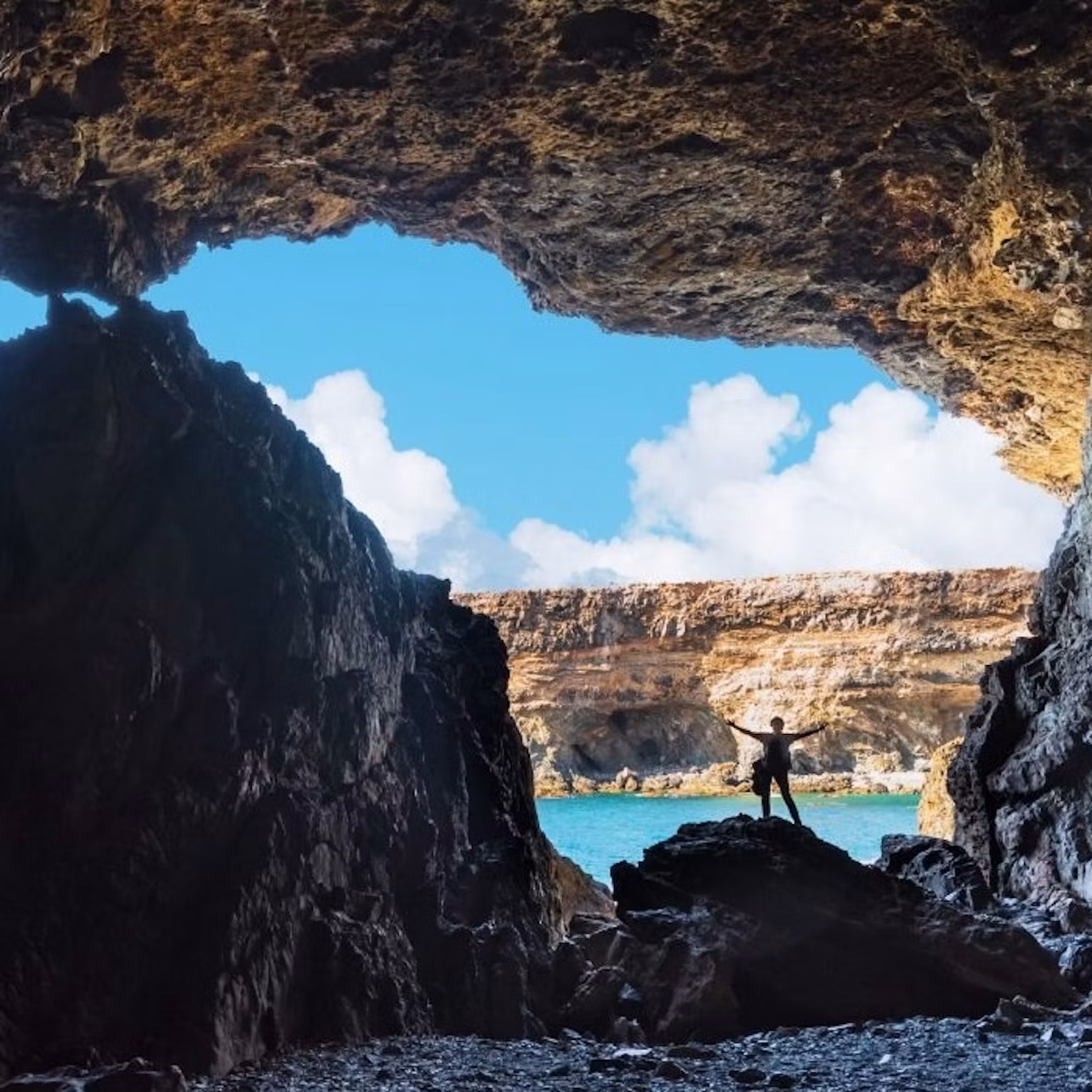 A person stands on rocks at the entrance of a cave overlooking a blue sky with clouds and a distant cliff.