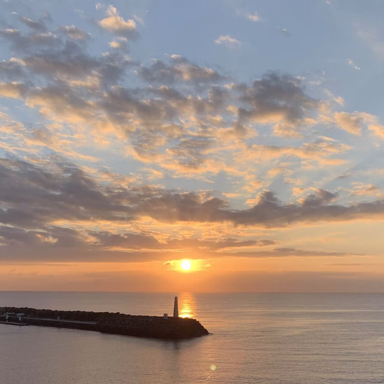 Sunset over calm ocean with a lighthouse on a rocky pier, under a sky filled with scattered clouds reflecting the sunset glow.