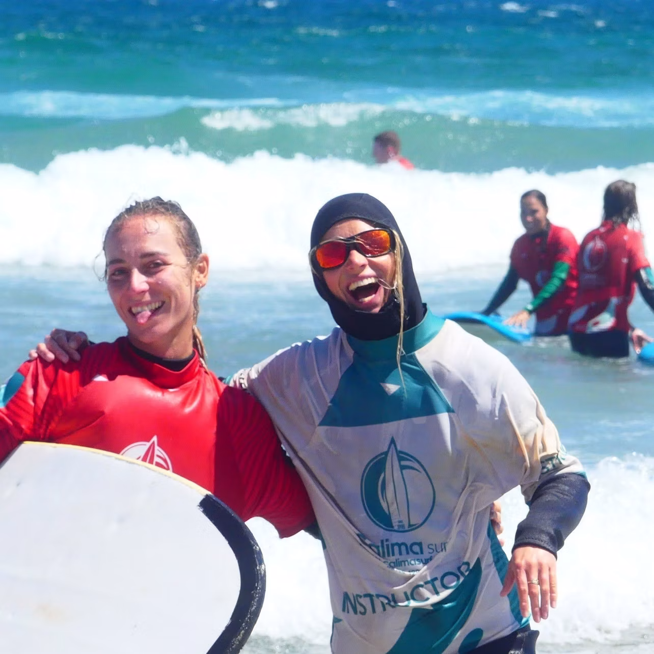 Two people in wetsuits smiling and posing with surfboards on a sunny beach, others surfing in the background.
