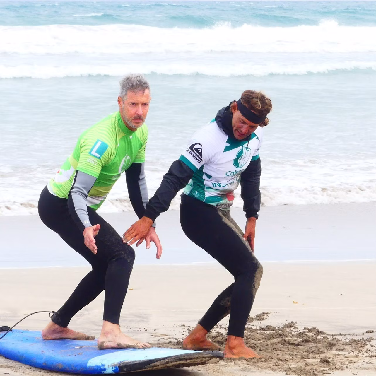 A man in a green shirt practices surfing poses on a blue surfboard on the beach while another man in a white shirt instructs him.