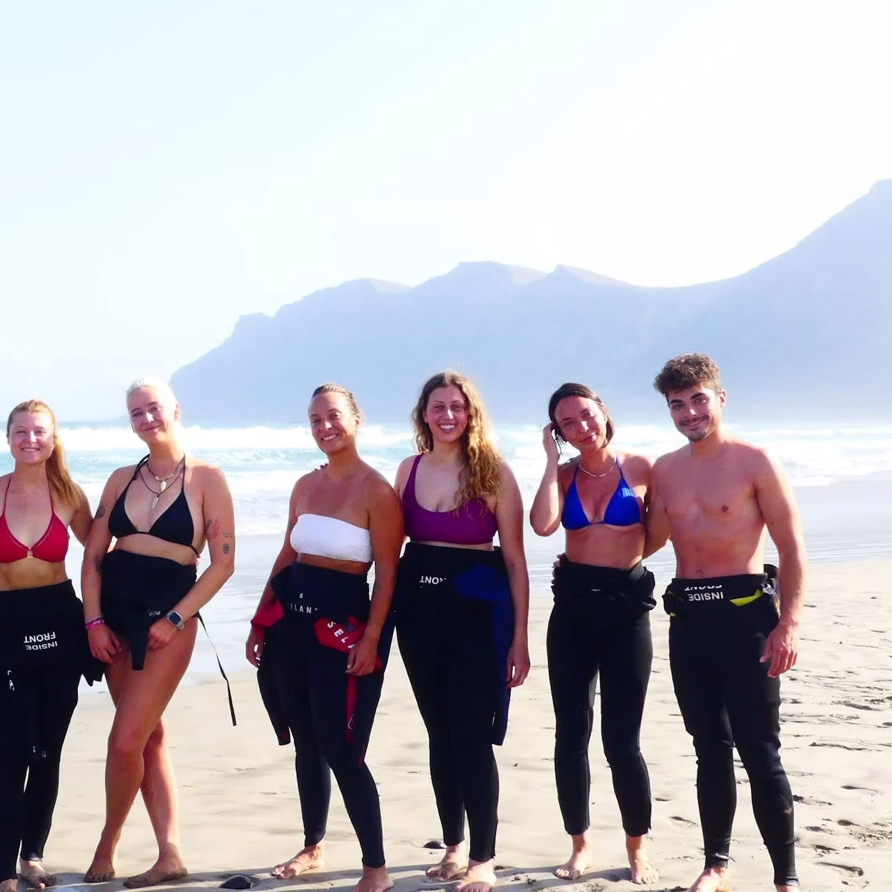 Six people in swimsuits and wetsuit pants stand on a beach with mountains in the background.