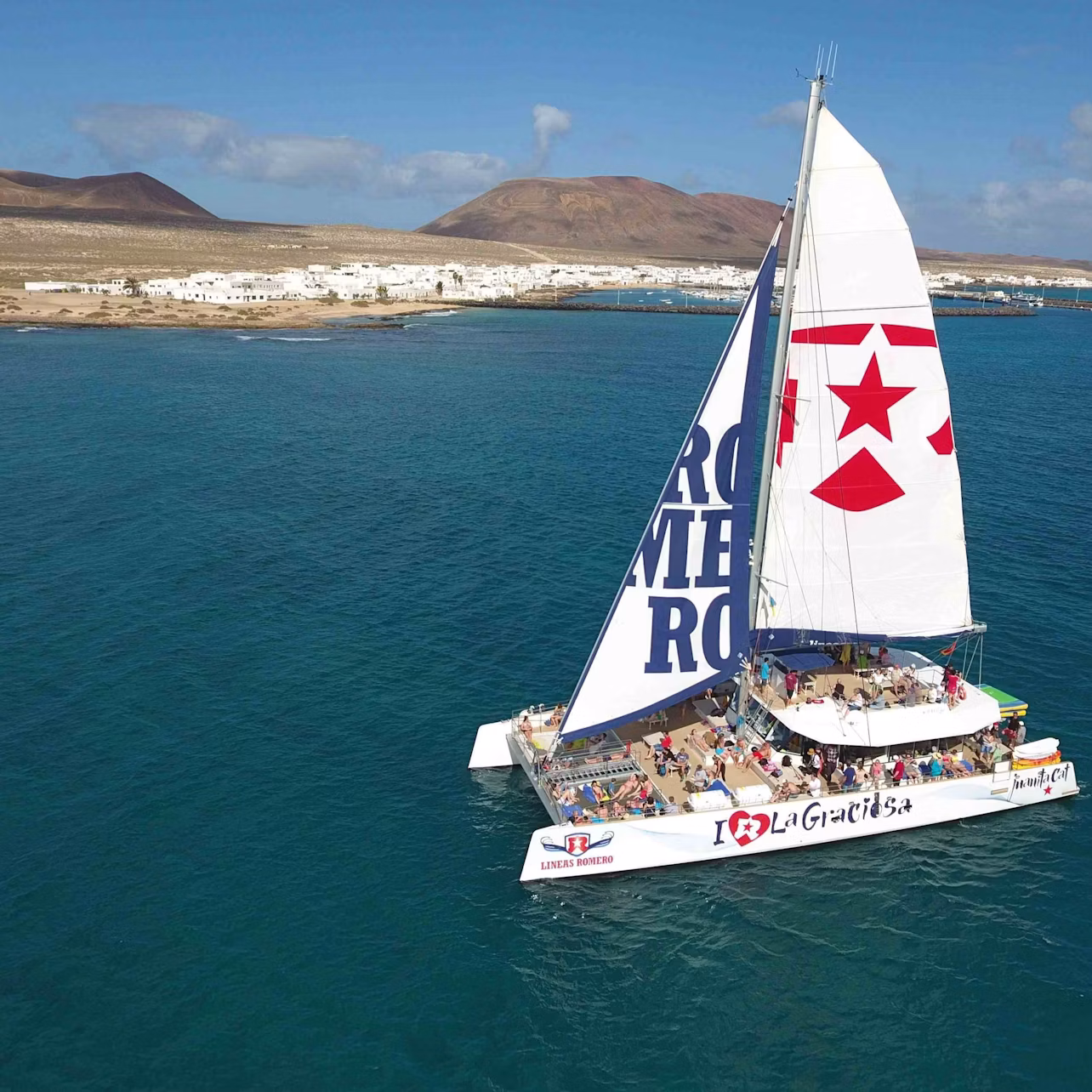 Catamaran with "I ❤️ La Graciosa" and star on sails, sailing in blue water near a coastal town with white buildings and hills.