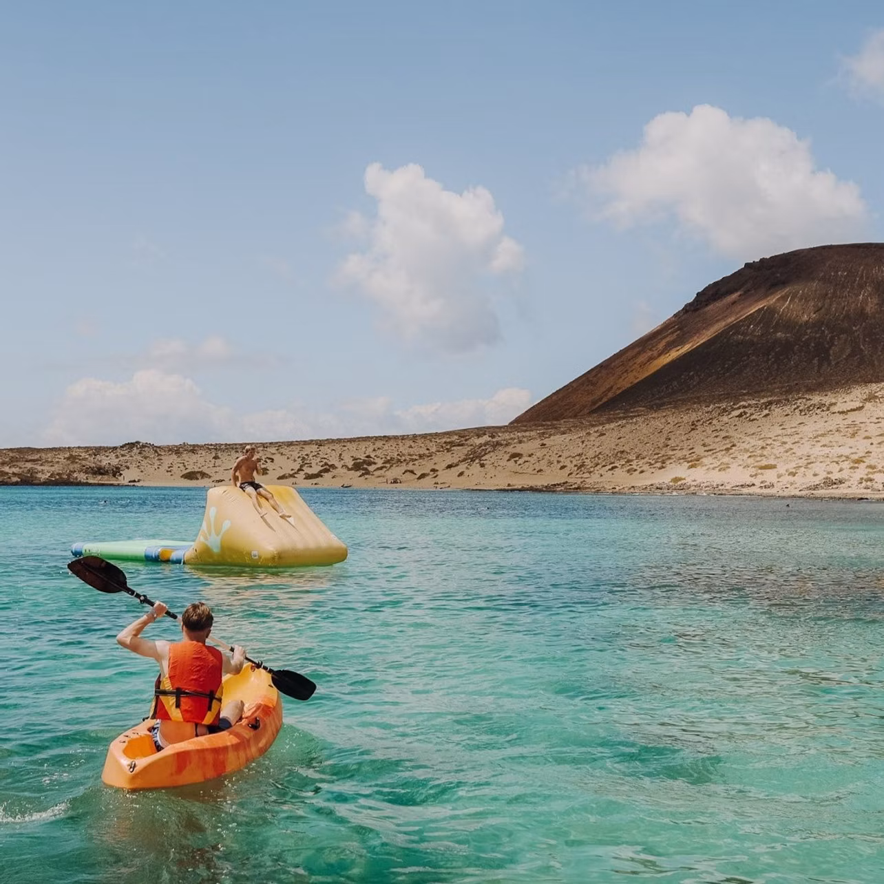 Person kayaking towards an inflatable structure on turquoise water, with a rocky hill and cloudy sky in the background.