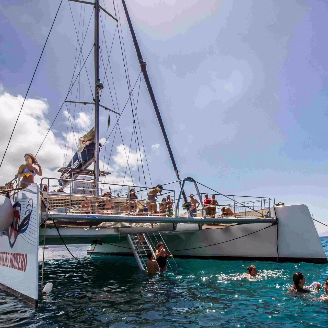 People enjoying a catamaran trip, swimming in clear blue water, with others lounging on the deck under a bright, sunny sky.