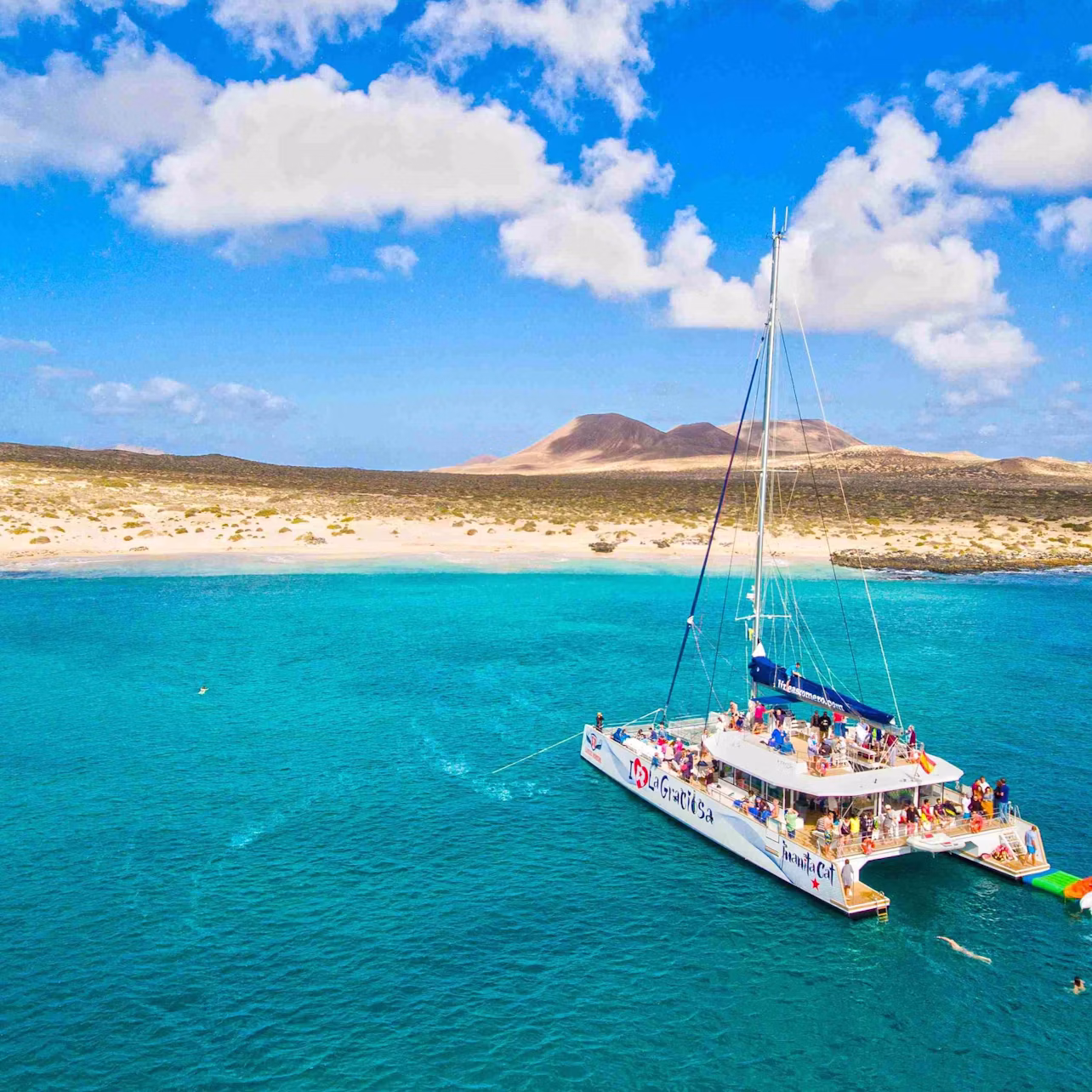 A catamaran with people onboard is anchored in clear blue waters near a sandy shore with mountains in the background under a blue sky.