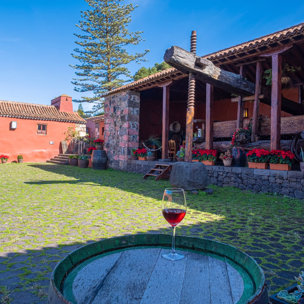 Glass of red wine on a wooden barrel in a courtyard with stone buildings, potted plants, and a traditional wine press.