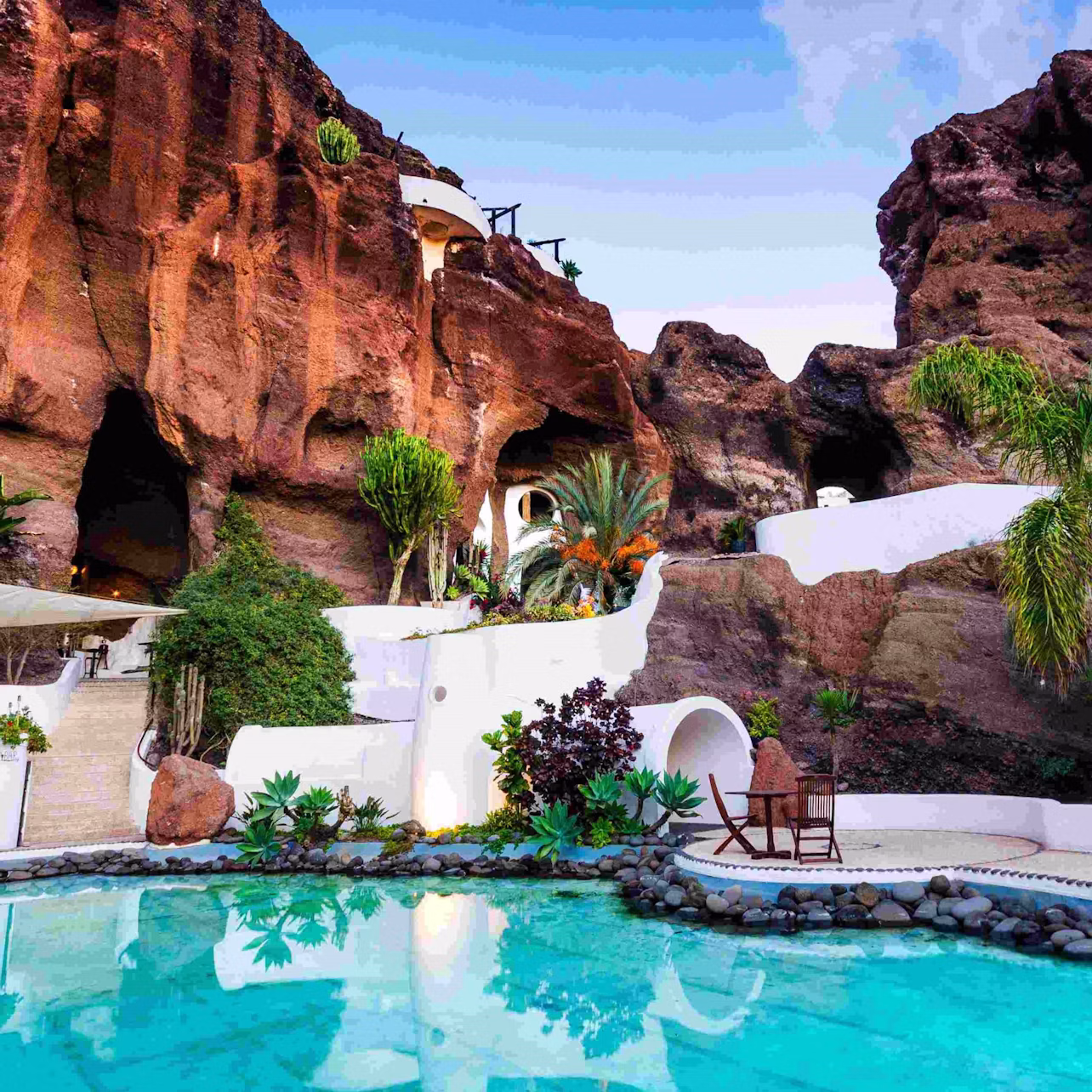 A serene pool with reflections of lush plants and white buildings carved into a rocky hillside under a partly cloudy sky.