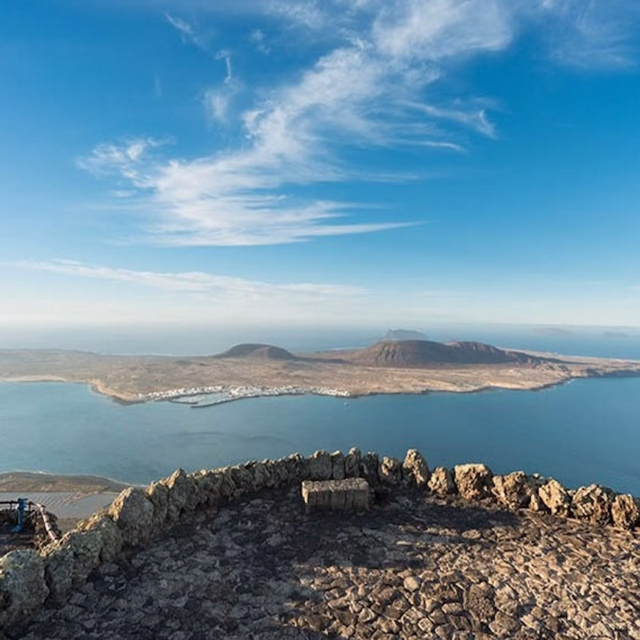 Rocky lookout point with a stone bench overlooking a coastline, distant mountains, and a clear, blue sky with wispy clouds.