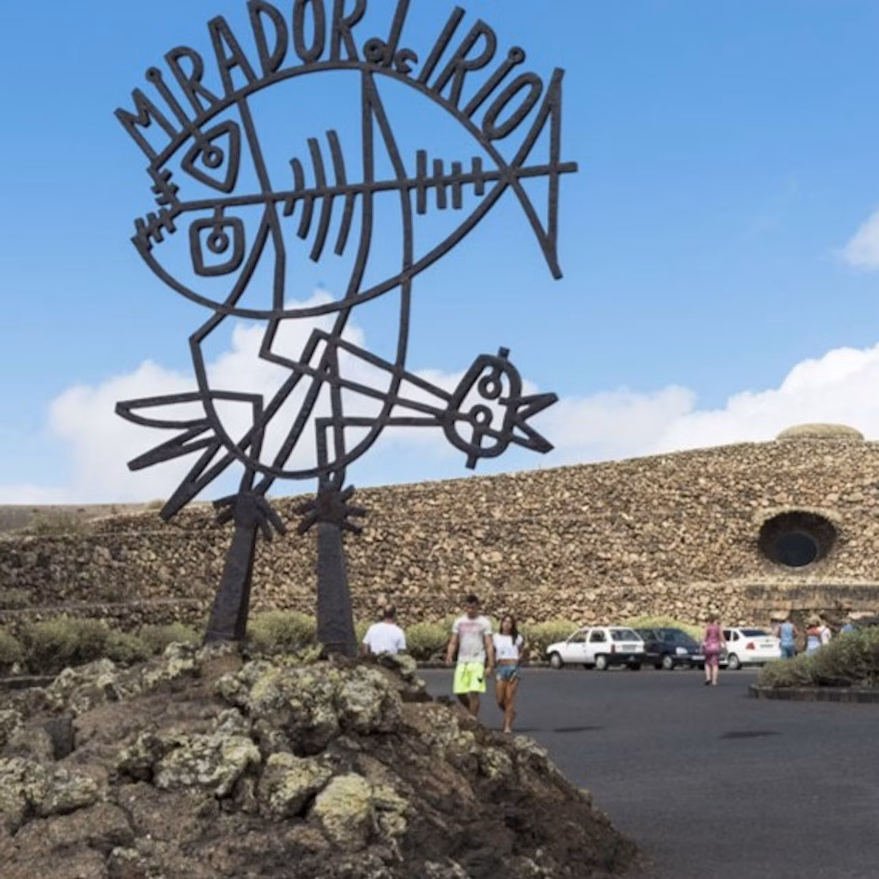 A metal sculpture reading "MIRADOR DEL RIO" stands in front of a stone building with a circular window. People and parked cars nearby.