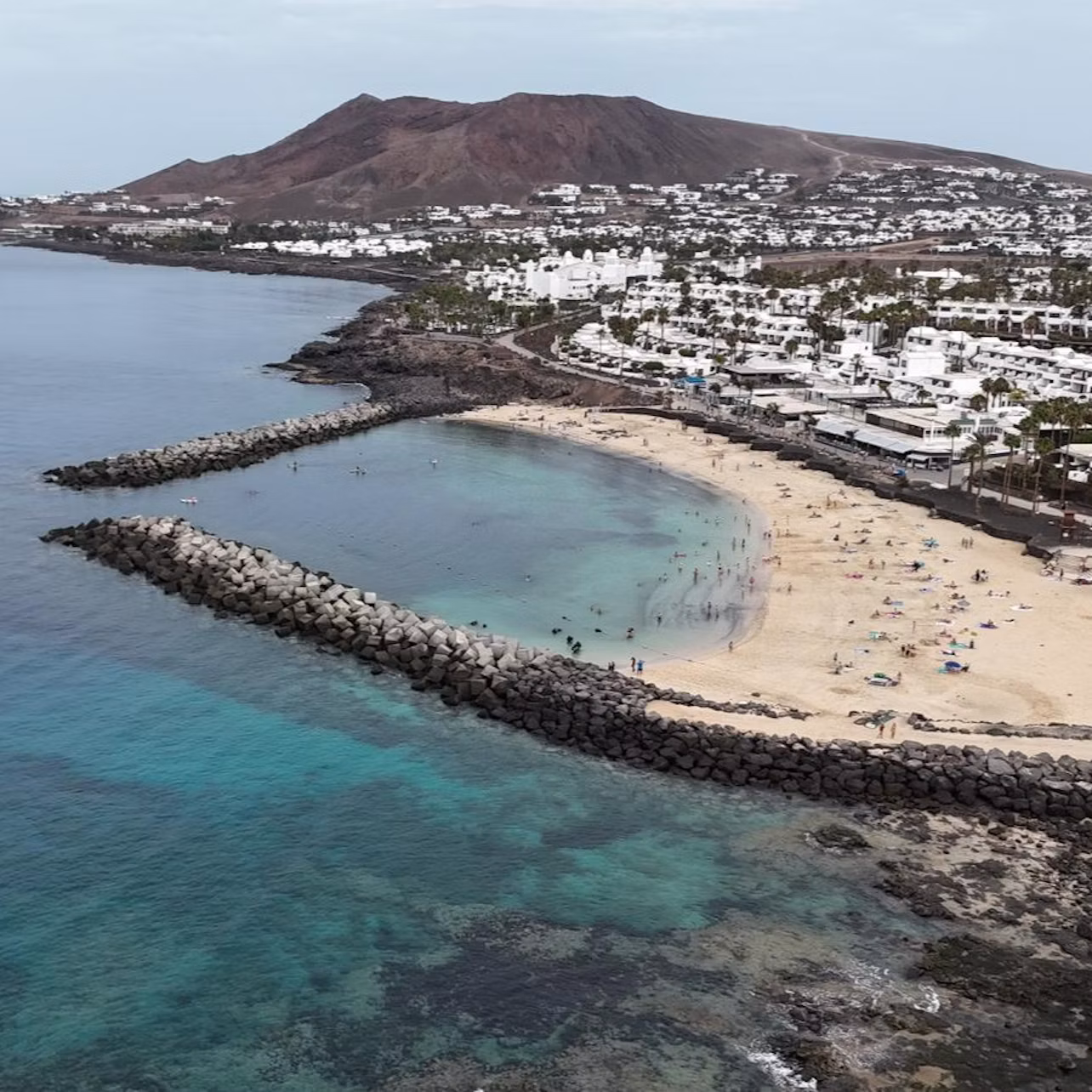 Aerial view of a beach with sand, clear water, people, and rock barriers. White buildings and a large hill are in the background.
