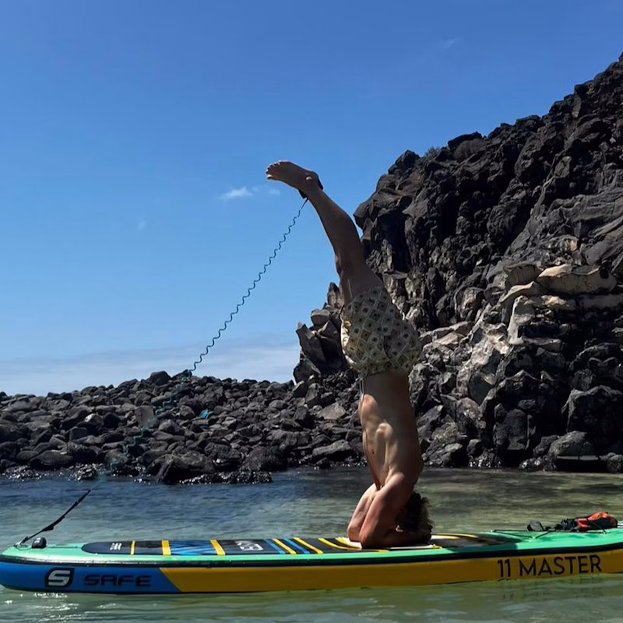 Person performing a headstand on a paddleboard in calm water near rocky shore under a clear blue sky.