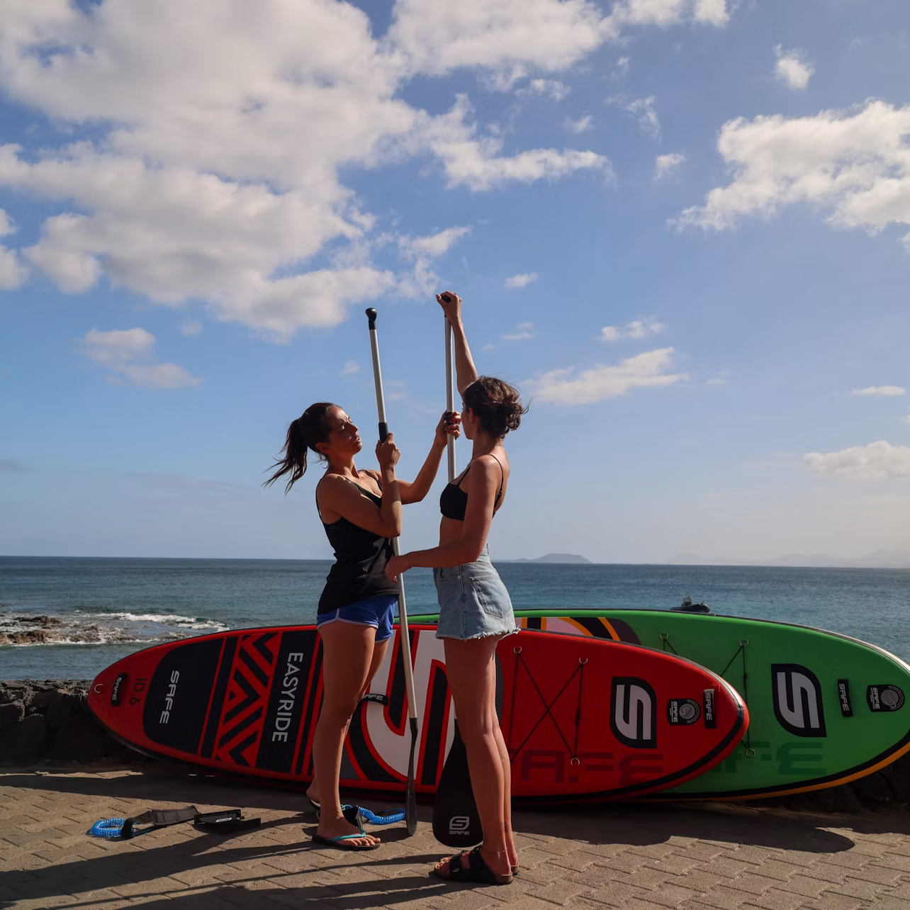 Two women standing on a paved path with paddleboards and paddles, adjusting paddle lengths near the ocean under a partly cloudy sky.