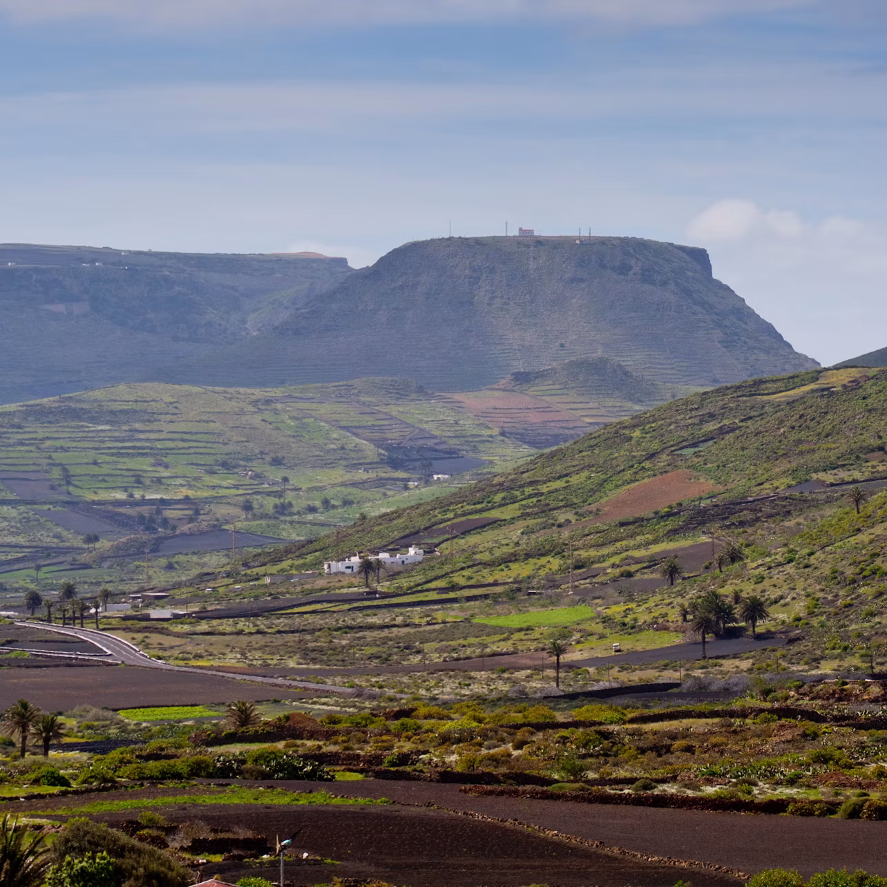 A lush green valley with terraced fields, scattered houses, and a flat-topped mountain under a partly cloudy sky.