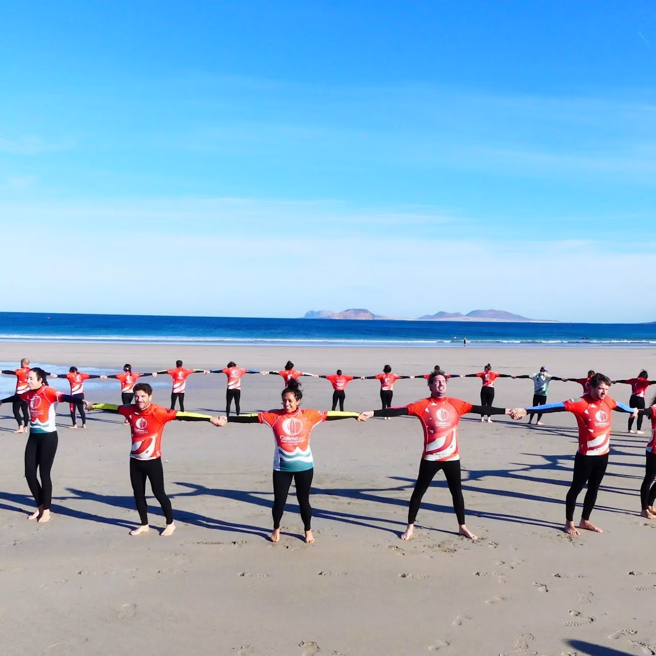 A group of people on a beach wearing red shirts and black pants, holding hands in a line under a clear blue sky.