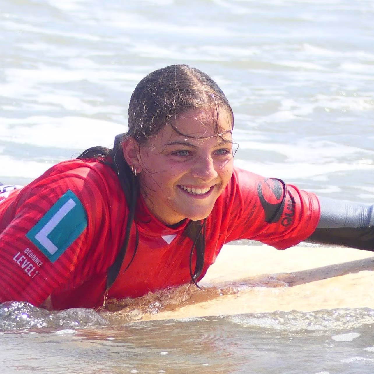 A person in a red wetsuit smiles while lying on a surfboard in shallow water.