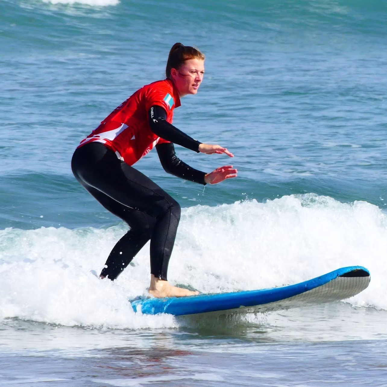 Person wearing a red shirt and black wetsuit surfing on a blue surfboard in the ocean waves.
