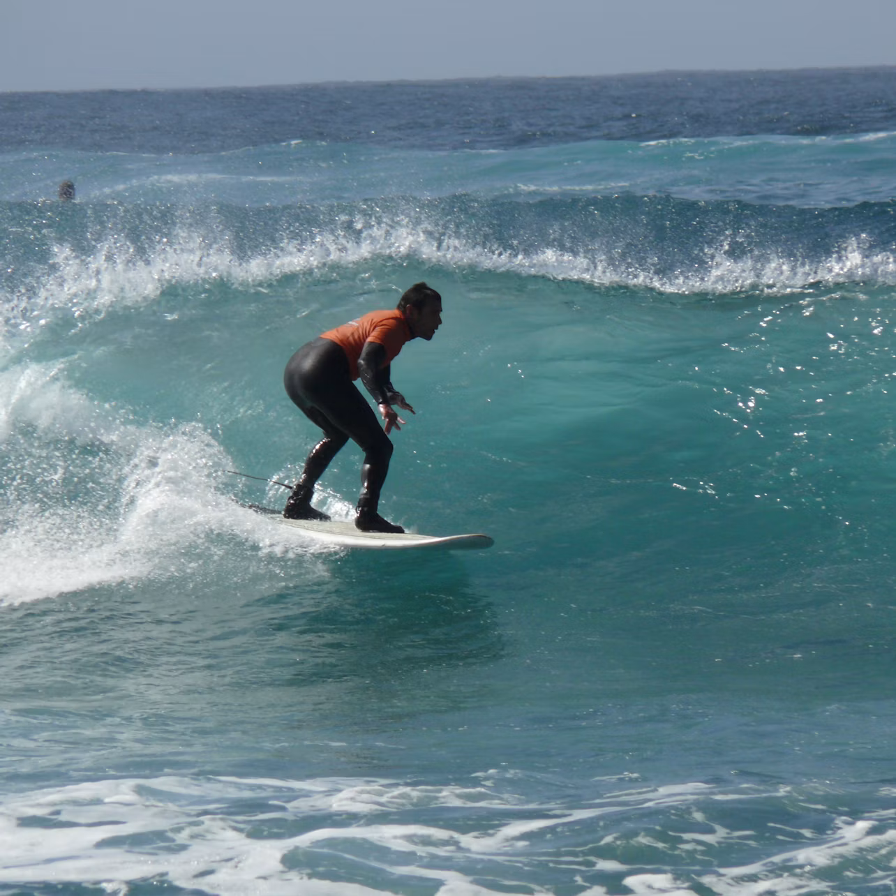 Person surfing on a large, blue wave in the ocean, wearing a wetsuit and an orange shirt, under clear skies.