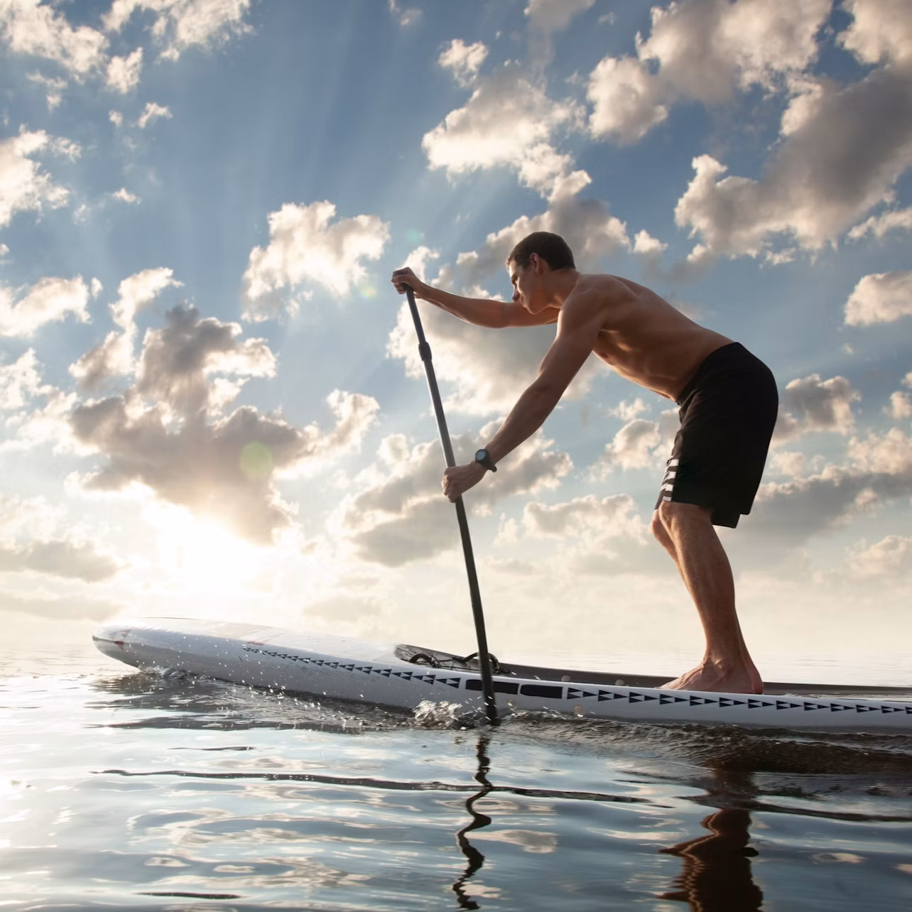 Person paddleboarding on a calm body of water at sunset, with a partly cloudy sky and sun rays visible.