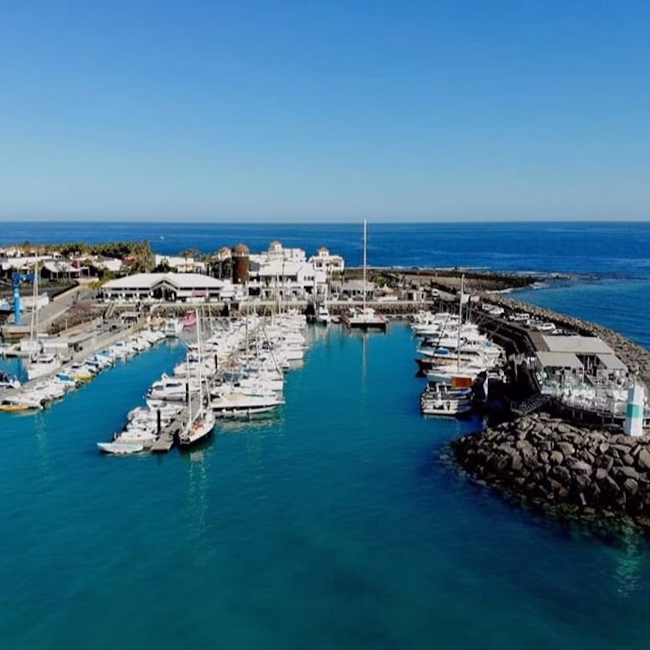Marina with numerous boats docked in clear blue water, surrounded by buildings and rocky breakwaters under a clear blue sky.