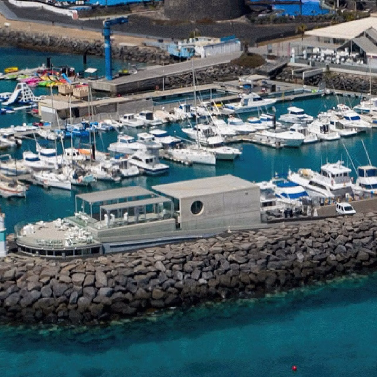 Aerial view of a marina filled with boats, adjacent to rocky breakwaters and a modern building near turquoise waters.