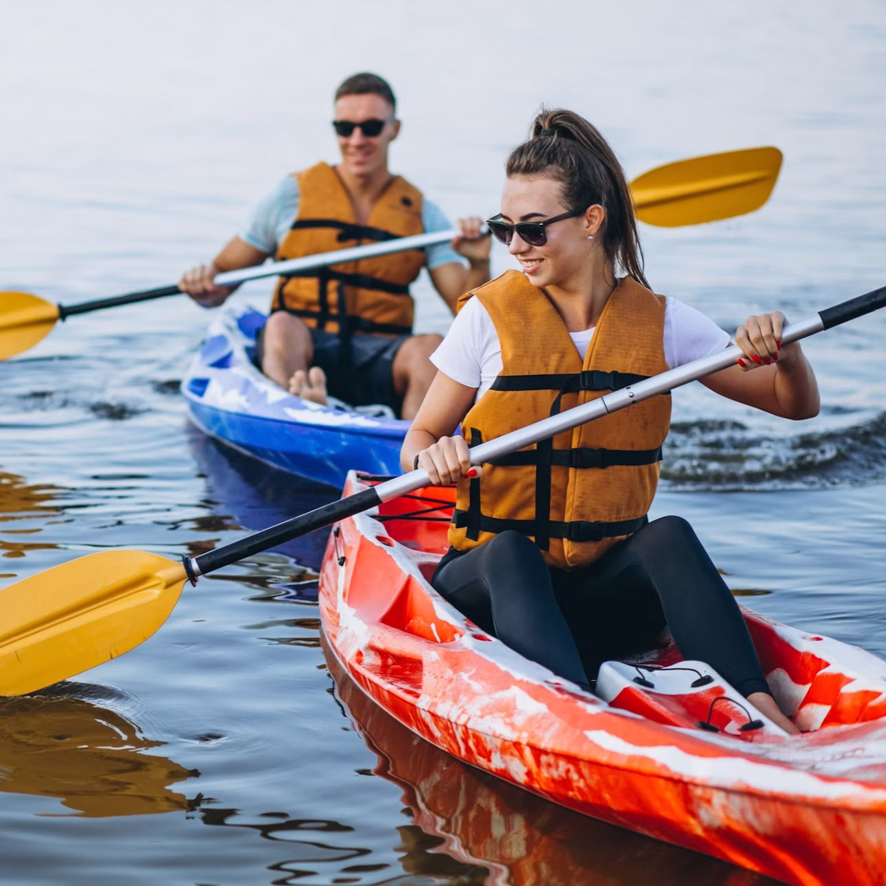 Two people wearing life vests and sunglasses paddling kayaks on calm water; one in a red kayak, the other in a blue kayak.
