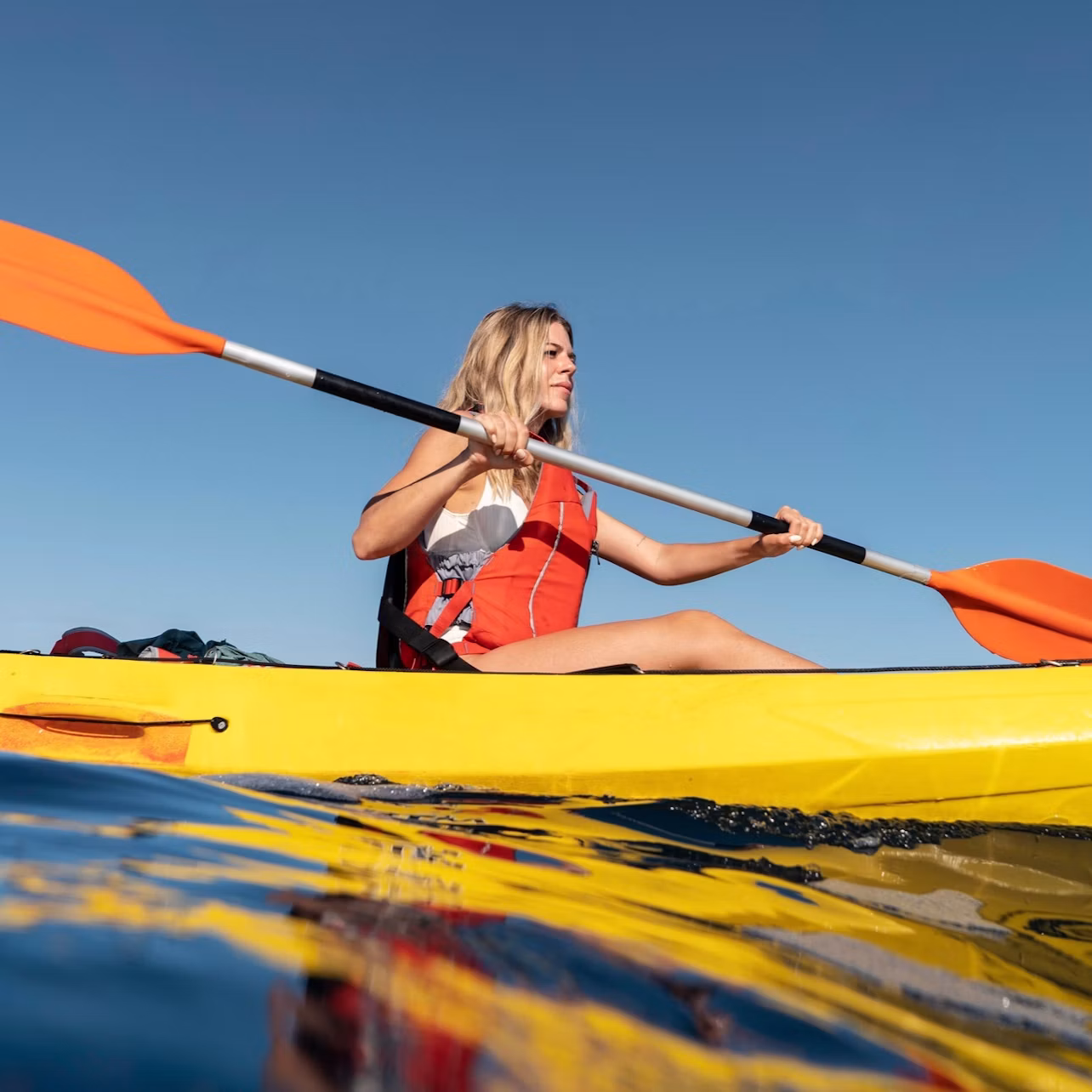 A person in a yellow kayak wearing a red life vest paddling on calm water under a clear blue sky.