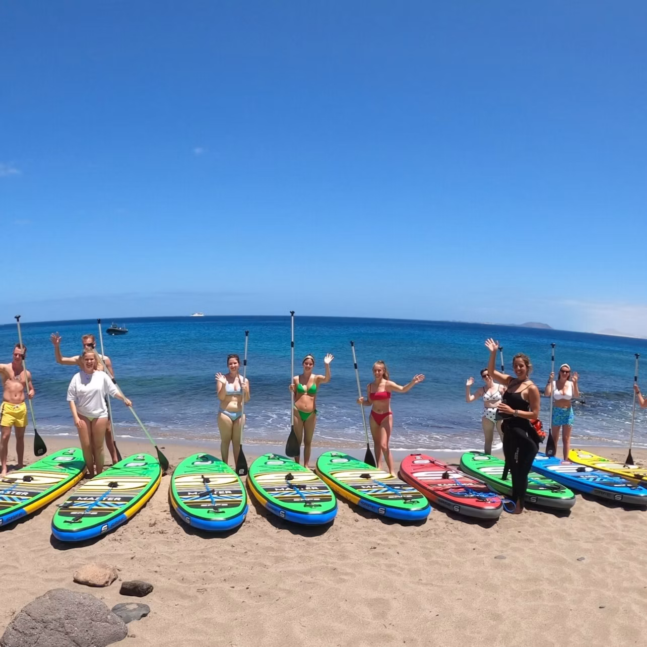 A group of people stands on a sandy beach holding paddles, with paddleboards laid out in front of them and the ocean in the background.