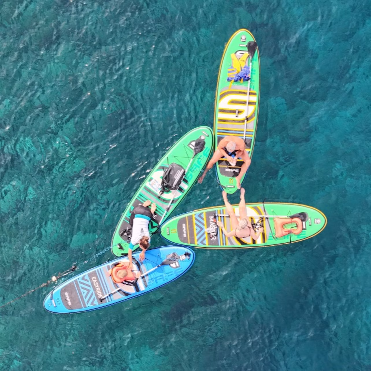 Four people relaxing on paddleboards clustered together on clear turquoise water.