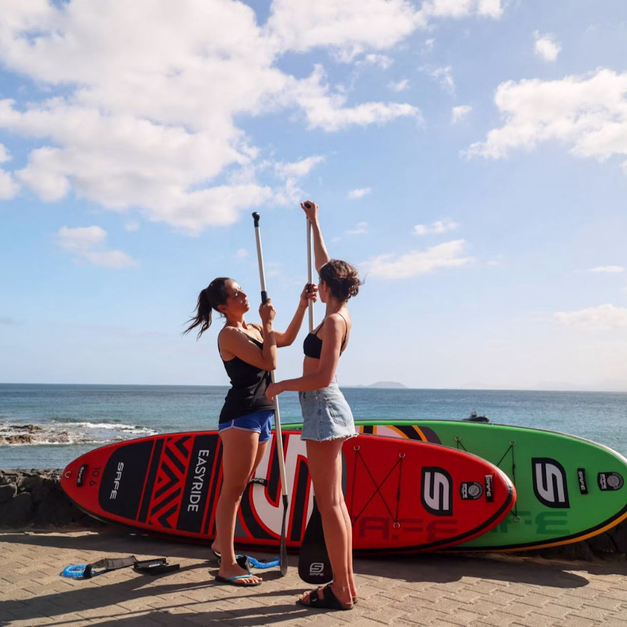 Two people stand on a beach holding paddles, with red and green stand-up paddleboards in the background under a blue sky.