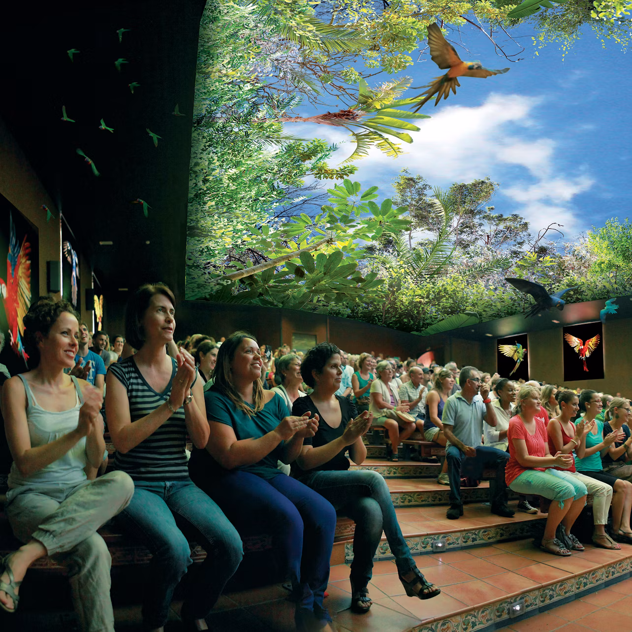 Audience clapping in an indoor theater with a ceiling designed to look like a jungle canopy, complete with birds and blue sky.