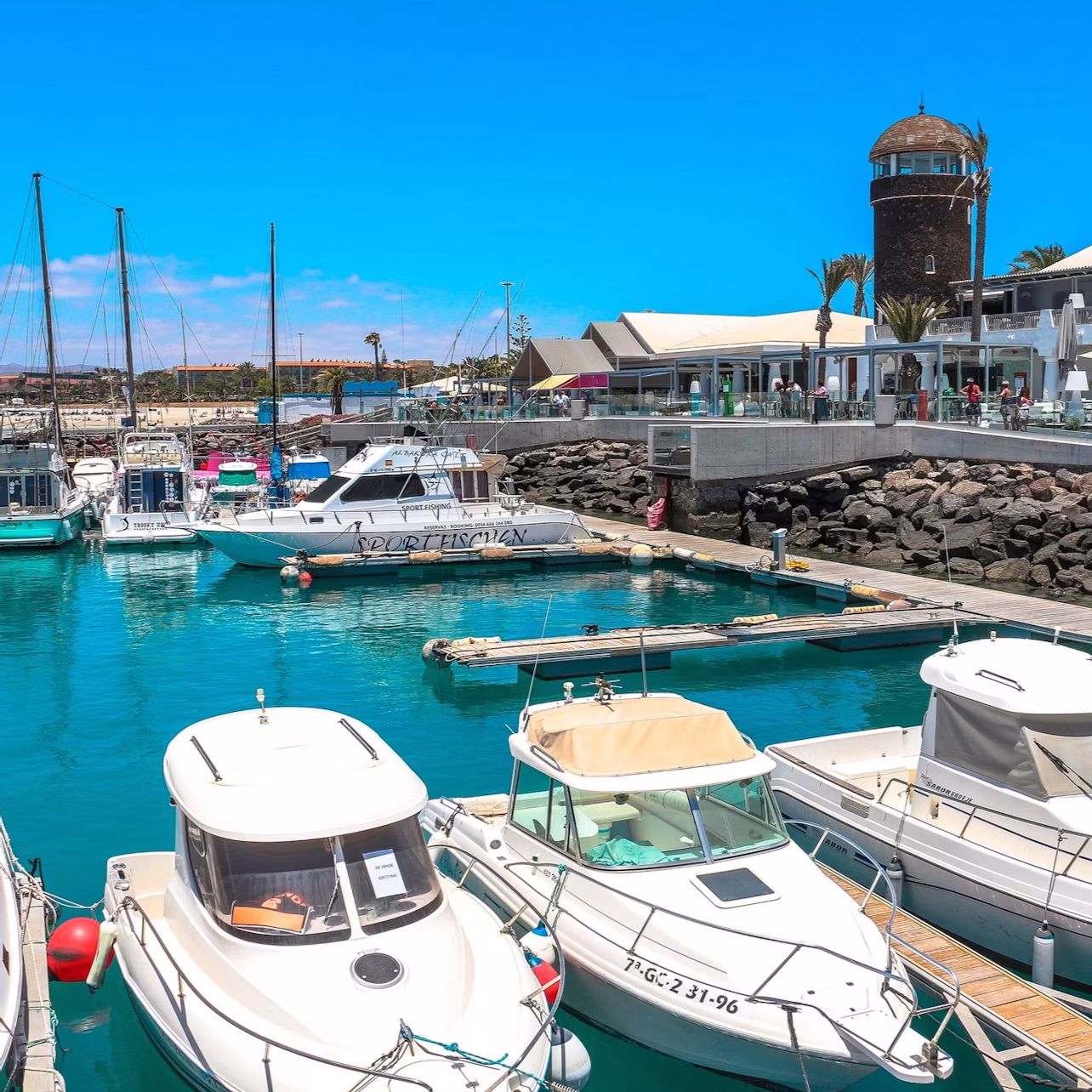 Docked boats in a marina with a stone building, shops, and a café in the background under a clear blue sky.