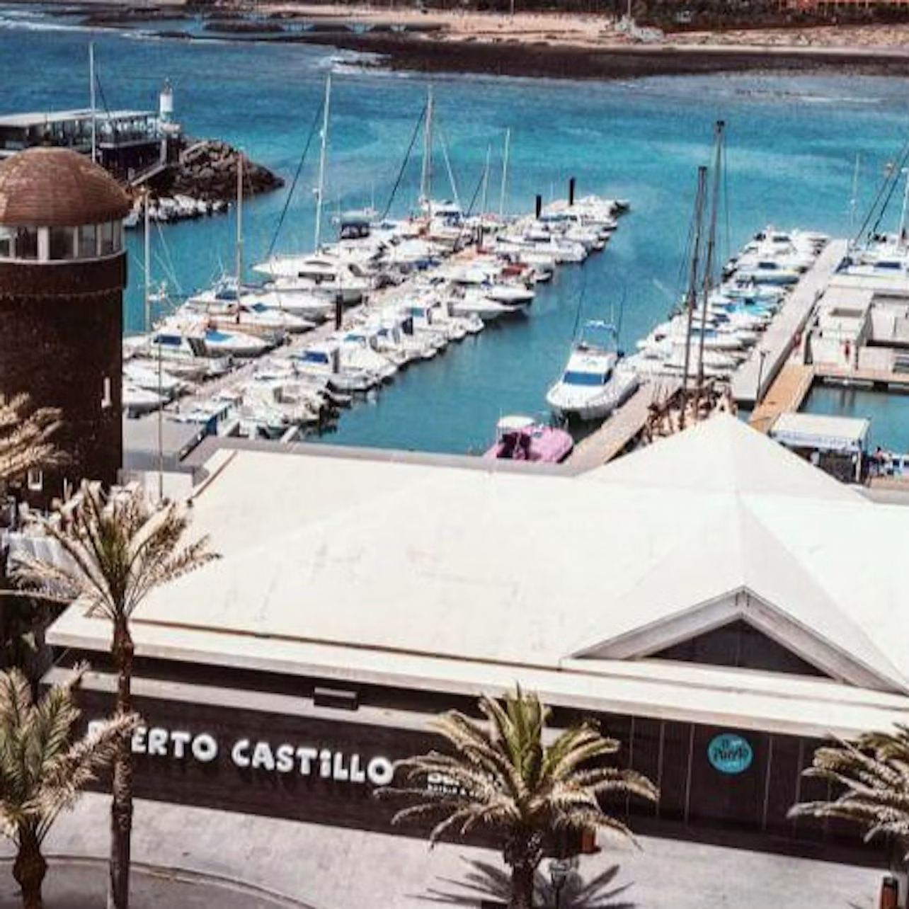 Marina with numerous docked boats, turquoise water, a cylindrical stone tower, and a building labeled "Puerto Castillo." Palm trees in foreground.